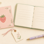 Pastel drawing of a strawberry greeting card, open mint journal, floral gift tag, and lavender pen.