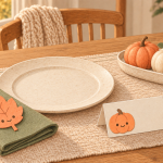 Cozy autumn table illustration with a smiling pumpkin place card, a leaf tag on a sage napkin, decorative pumpkins and a knitted throw by the window.