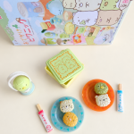 Sumikkogurashi miniature rice-ball characters on blue and orange plates, a tiny lunchbox, bottle and chopsticks with their packaging.