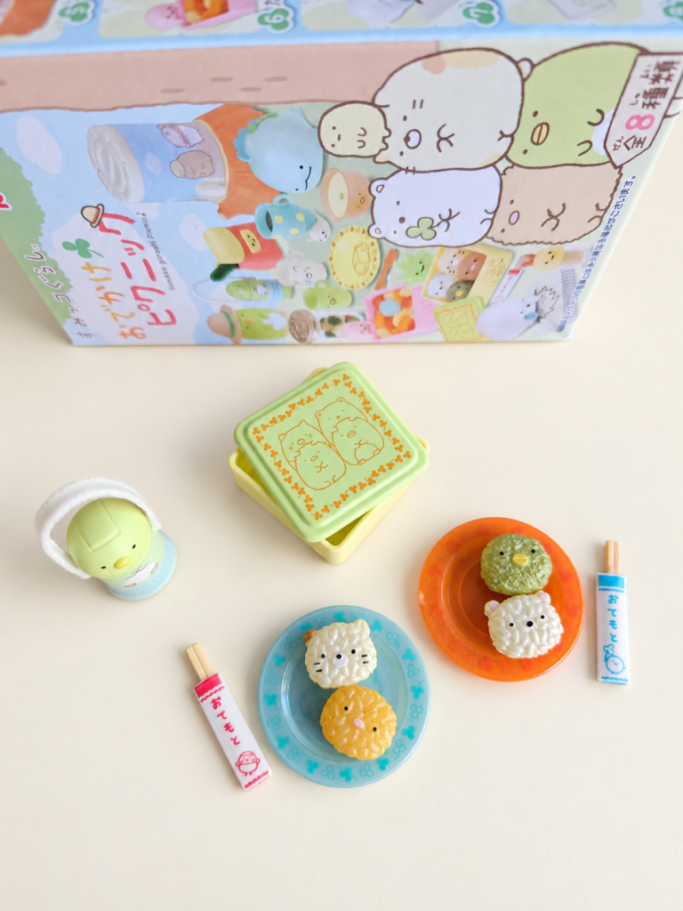 Sumikkogurashi miniature rice-ball characters on blue and orange plates, a tiny lunchbox, bottle and chopsticks with their packaging.